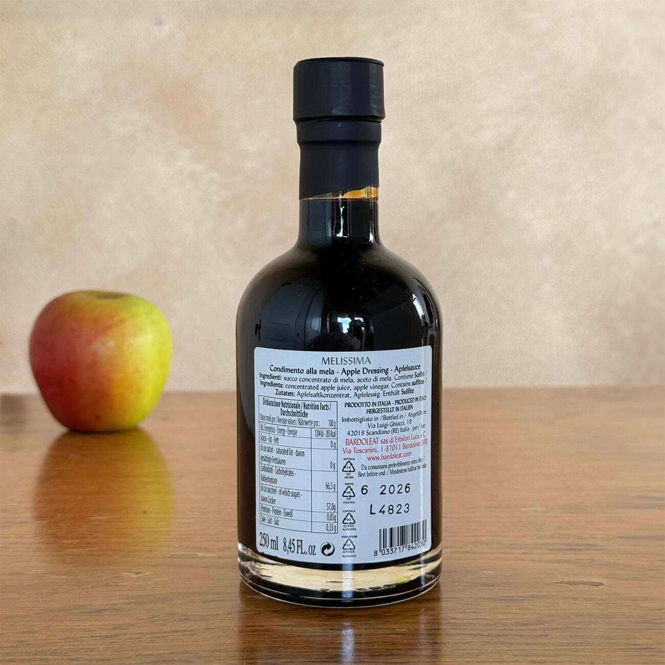 Die Flasche Balsamico ist von hinten auf einem Holztisch zu sehen. Im Hintergrund liegt verschwommen erkennbar ein Apfel.