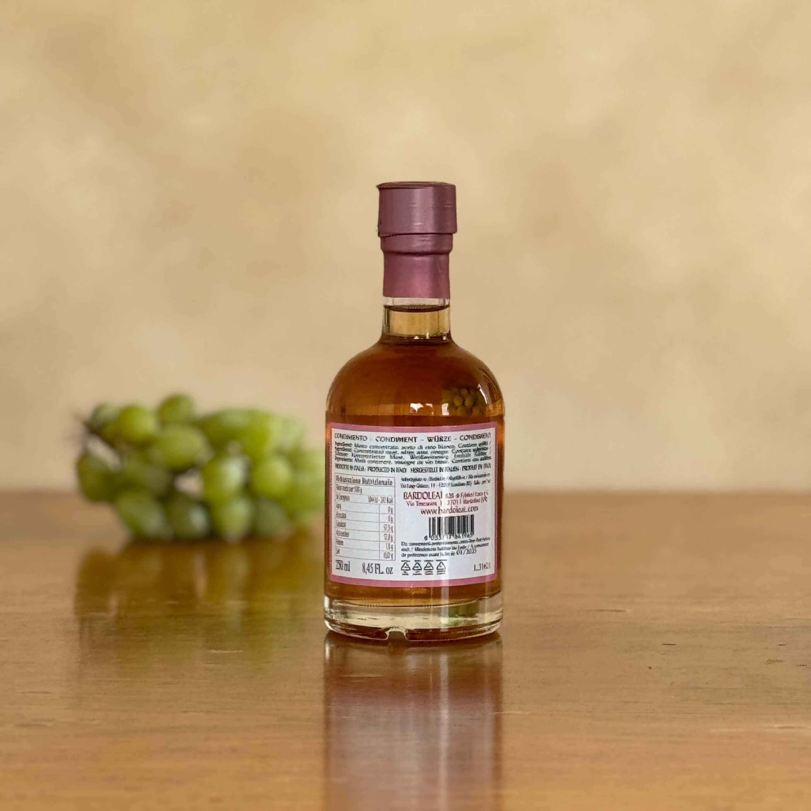 Eine Flasche Condimento Rosé steht auf einem Holztisch und ist von hinten zu sehen. Im Hintergrund liegen verschwommen erkennbar Weintrauben.
