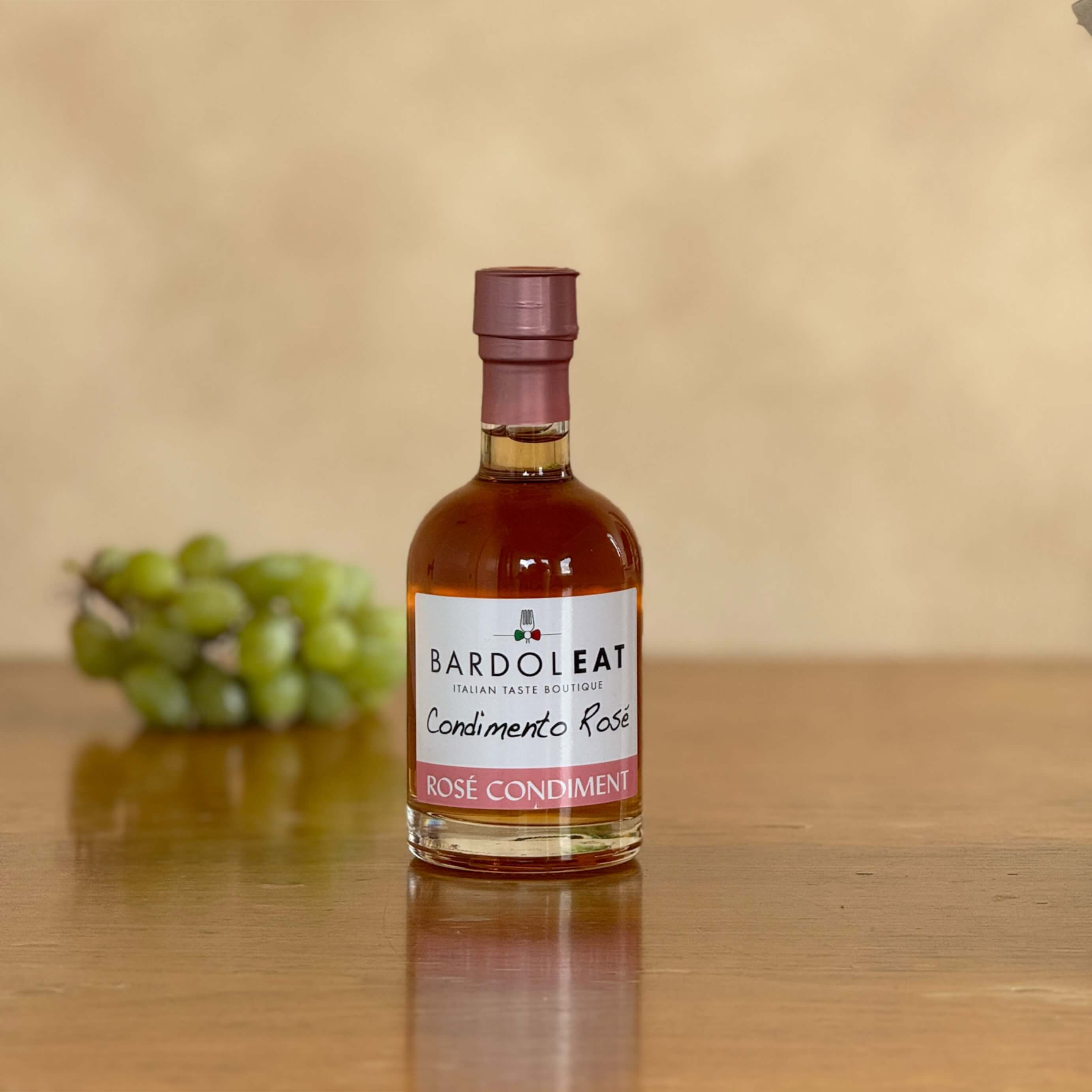 Eine Flasche Condimento Rosé steht auf einem Holztisch und ist von vorne zu sehen. Im Hintergrund liegen verschwommen erkennbar Weintrauben.